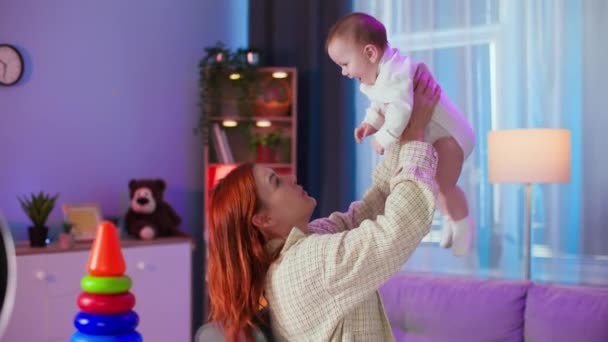 μητρότητα, ευτυχισμένη νεαρή γονιός ρίχνει την κόρη της ψηλά και την πιάνει με τα χέρια στο άνετο δωμάτιο - Πλάνα, βίντεο