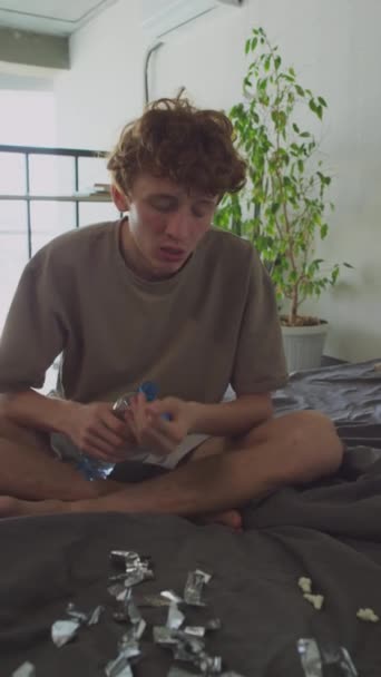 Portrait vertical d'un jeune homme affamé assis sur un lit non fait avec des mouchetures dessus, buvant de l'eau de la bouteille et regardant la caméra tout en ressentant de la fatigue le matin après la fête - Séquence, vidéo