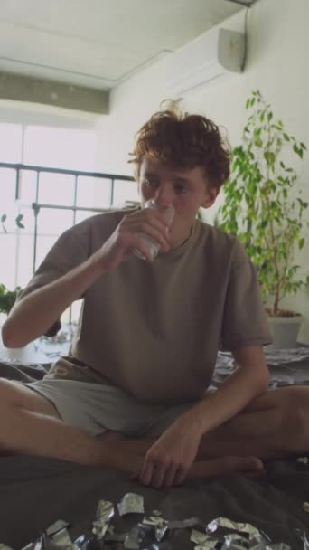 Vertikale Aufnahme des hungrigen Kerls, der morgens nach der Party auf dem Bett sitzt und Wasser mit aufgelöster Brausetablette trinkt - Filmmaterial, Video