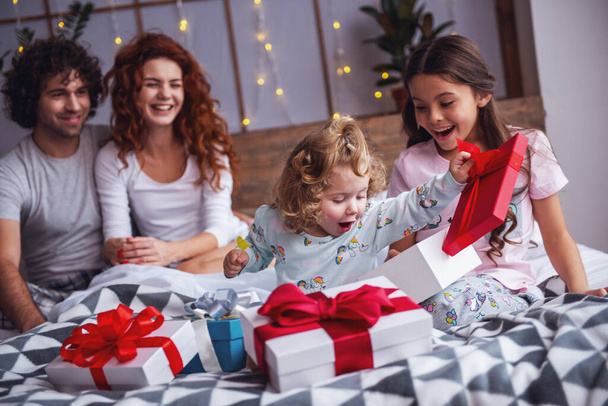 それはクリスマスの時間です!幸せな家族のベッドに座って、両親が笑みを浮かべながら自分の娘がプレゼントを開いています。 - 写真・画像