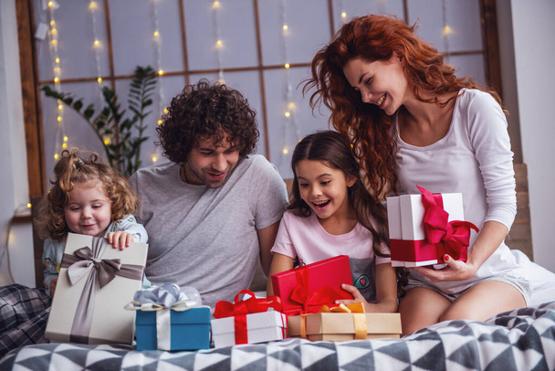 It's Christmas time! Happy family sitting in bed, parents are smiling while their daughters are opening the presents - Photo, Image