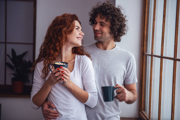 Para piękny jest picie gorącego kakao z prawoślazu, patrząc na siebie i uśmiechając się podczas odpoczynku w domu - Zdjęcie, obraz