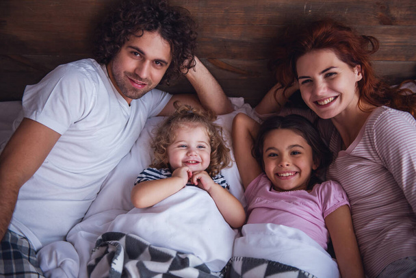 Счастливые родители и их милые дочери смотрят в камеру и улыбаются, лежа вместе в постели дома
 - Фото, изображение