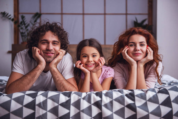 Gelukkige ouders en hun dochter kijken naar de camera en lachen terwijl ze thuis op het bed liggen. - Foto, afbeelding