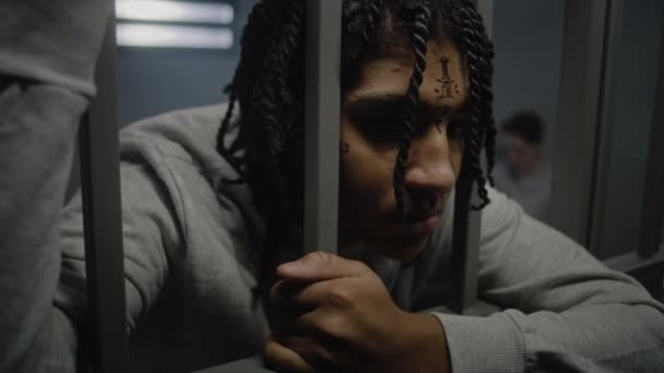 Close up shot of angry African American teenage prisoner with face tattoos standing in prison cell, holding metal bars. Young inmate serves imprisonment term for crime in jail. Youth detention center. - Footage, Video