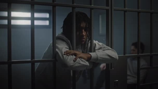 Depressed African American teenager with tattoos stands in prison cell in jail and looks at camera. Young inmate serves imprisonment term for crime. Juvenile detention center or correctional facility. - Footage, Video