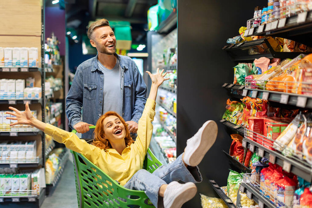 Gioioso coppia avendo divertimento mentre l'acquisto di cibo in supermercato, felice uomo spingendo carrello della spesa con la moglie seduta dentro - Foto, immagini