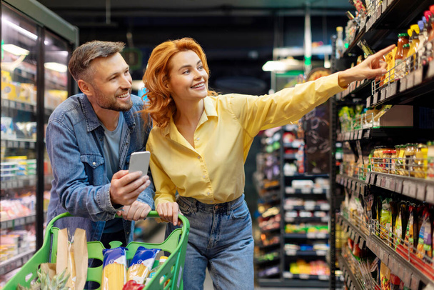 Couple faisant des courses ensemble, homme utilisant un téléphone portable, femme pointant du doigt une étagère, choisissant des produits alimentaires dans un supermarché - Photo, image