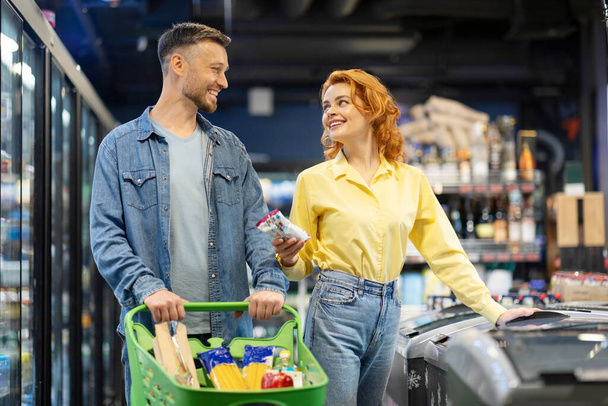 Ευτυχισμένο νεαρό ζευγάρι ψωνίζει μαζί στο σούπερ μάρκετ, σύζυγος και γυναίκα στέκεται δίπλα στον καταψύκτη με τρόλεϊ γεμάτο τρόφιμα - Φωτογραφία, εικόνα