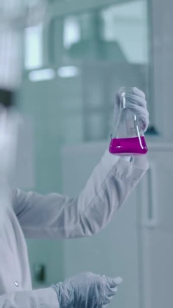 Mediana vertical de joven investigadora asiática en máscara facial de seguridad, gafas, bata de laboratorio sosteniendo y examinando frasco con solución química púrpura, buscando residuos - Metraje, vídeo