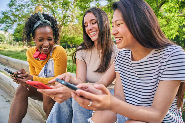 Drei junge Frauen teilen glückliche Momente auf einer Bank und lächeln in ihre Handys. Sie sitzen unter einem Baum und genießen Freizeit und Freundschaft in einer lustigen und erholsamen Adaption moderner Technologie - Foto, Bild