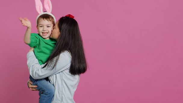 Mother lifting up her little boy and kissing him in studio, showing love and hugging cute kid against pink background. Sweet mom and son embracing each other and laughing. Camera A. - Photo, Image