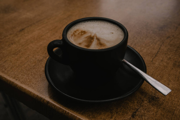 Een zwarte koffiebeker, mok staat op een oude houten tafel. Aromatische smakelijke cappuccino of cacao in een café. Aromatische en warme ochtenddrank in cafetaria.  - Foto, afbeelding