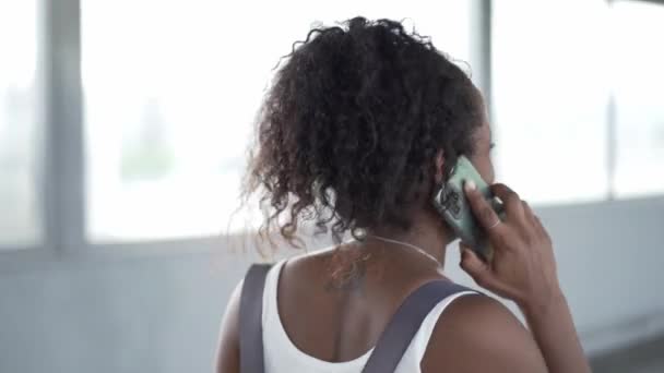 Вид ззаду на невпізнаваний молодий латиноамериканський турист, який розмовляє по мобільному телефону під час прогулянки по коридору аеропорту або транспортній станції. Африканська жінка відповідає на мобільний дзвінок. Люди, які подорожують у відпустку - Кадри, відео