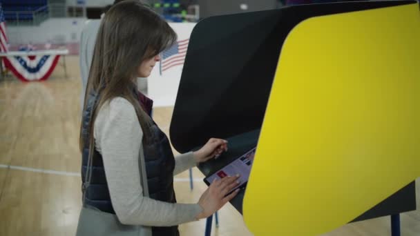 ZAPORIZHYA, UCRÂNIA - MARÇO 15, 2024: Mulher caucasiana, eleitor pensa e faz a escolha de pé perto da cabine de votação na estação de votação. Cidadão dos EUA usa tablet para votar. Presidencial - Filmagem, Vídeo
