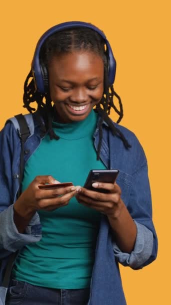 Вертикальне відео Посміхнена жінка додає метод оплати на сайті, насолоджуючись періодом продажів, студійним фоном. Дівчина робить покупки в Інтернеті, вводить інформацію про кредитну картку на смартфон, слухає музику - Кадри, відео