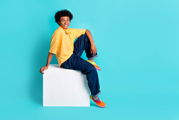 Full length photo of cool guy dressed yellow t-shirt jeans sit on cube near logo empty space isolated on turquoise color background. - Foto, Imagen