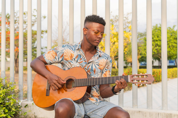 Skupiony Afrykanin w okularach przeciwsłonecznych i kwiatowej koszuli gra na gitarze, otoczony tętniącym życiem ogrodem i nowoczesną architekturą. - Zdjęcie, obraz
