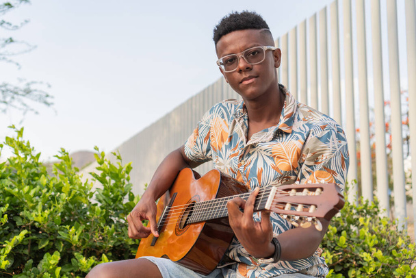 Selbstbewusster junger afrikanischer Mann mit Brille und floralem Hemd, der draußen, umgeben von grünem Laub, eine akustische Gitarre spielt. - Foto, Bild