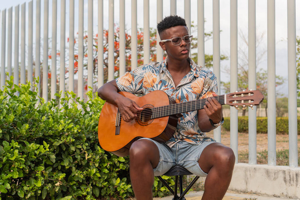 Un giovane concentrato che indossa una camicia tropicale strumenta una chitarra acustica in un ambiente sereno all'aperto con vegetazione lussureggiante e architettura moderna. - Foto, immagini