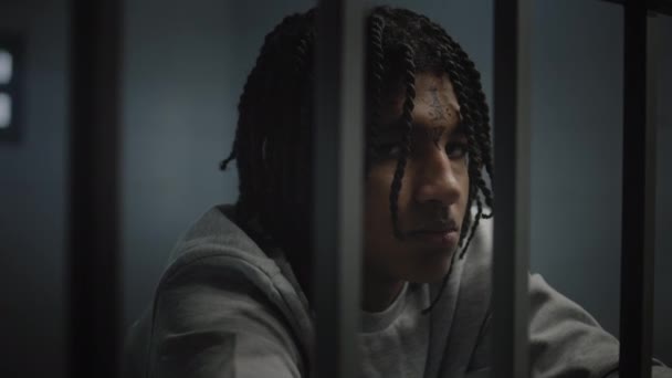 Yüzünde dövmeler olan kızgın Afrikalı Amerikalı genç mahkum hapisteki hücrede duruyor ve kameraya bakıyor. Genç suçlu, suçtan hapis cezasına çarptırıldı. Islahevi. Portre. - Video, Çekim