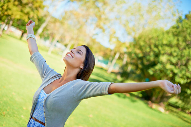Happy woman, freedom and nature for outdoor wellness, travel and fresh air in environment and summer. A young and casual person wit eyes closed and stretching arms for celebration and excited in park. - Photo, Image