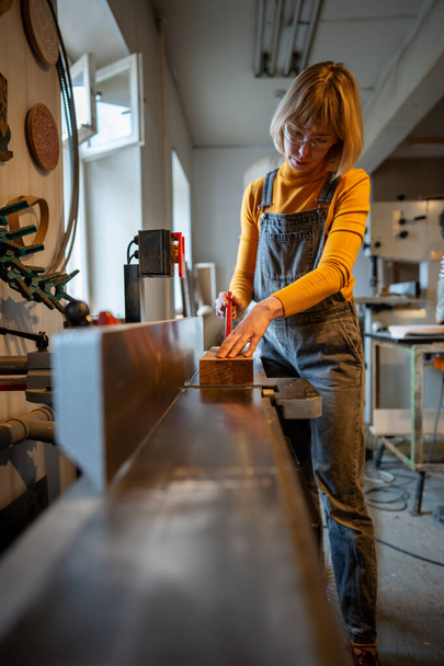 Προσεκτικά εστιασμένη γυναίκα ξυλουργός εργάζεται σε ηλεκτρική πλάνη φορώντας γυαλιά ασφαλείας. Συμπυκνωμένο γυναικείο joiner αλέθει ξύλινο μπλοκ για χειροποίητα ξύλινα έπιπλα. Εξοπλισμός για ξυλουργικές μικρές επιχειρήσεις. - Φωτογραφία, εικόνα