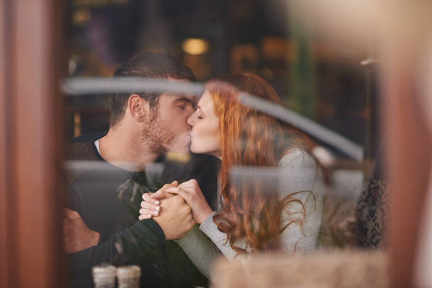 Couple, kissing and holding hands on date at cafe for bonding, romance and healthy relationship with relax. Man, woman and affection at coffee shop with lens flare, trust and love on anniversary trip. - Photo, Image