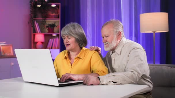 pensionati utilizzano la tecnologia moderna, felice vecchia donna con marito divertirsi giocando ai giochi per computer mentre seduto a tavola in una stanza accogliente - Filmati, video