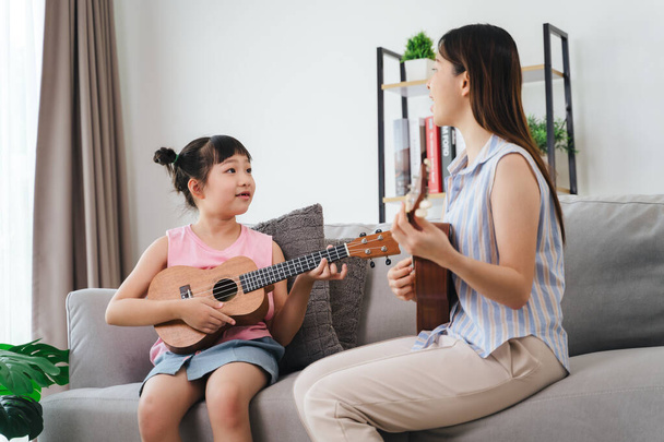 Una donna e un bambino sono seduti su un divano, con la donna che suona un ukulele. La bambina la sta guardando giocare. - Foto, immagini