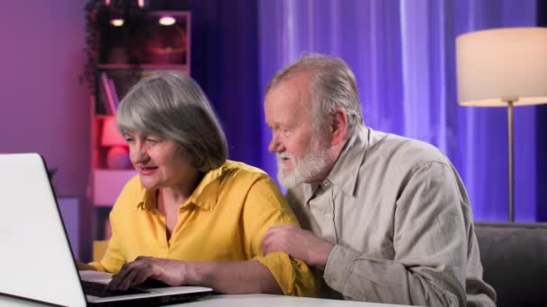 homem velho alegre e mulher se divertindo jogando jogos de computador modernos em um laptop on-line enquanto sentado à mesa em um quarto acolhedor - Filmagem, Vídeo