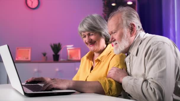 χαρούμενο ηλικιωμένο παντρεμένο ζευγάρι που διασκεδάζει παίζοντας παιχνίδια στον υπολογιστή σε ένα φορητό υπολογιστή στο τραπέζι σε ένα άνετο σαλόνι - Πλάνα, βίντεο