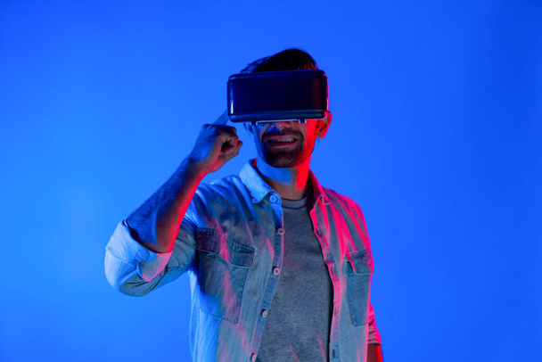 Man draagt virtual reality headset tijdens het invoeren in metaverse. Blanke man staan op kleurrijke neon licht en met behulp van futuristische digitale technologie virtuele bril of VR voor het spelen van games. Afwijking. - Foto, afbeelding