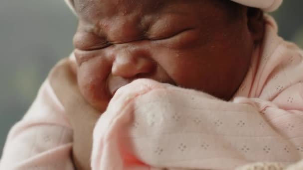 Closeup of African American infant girl in baby cap and light pink jumpsuit with mittens crying in mothers arms - Footage, Video