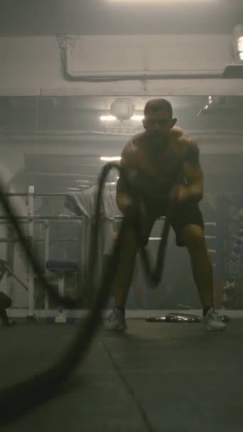 Profesjonalny bokser męski wykonuje trening cardio lub wytrzymałościowy przed walką o mistrzostwo. Atletyczny mężczyzna ćwiczy na linach bojowych w ciemnej siłowni bokserskiej. Strzał pionowy - Materiał filmowy, wideo
