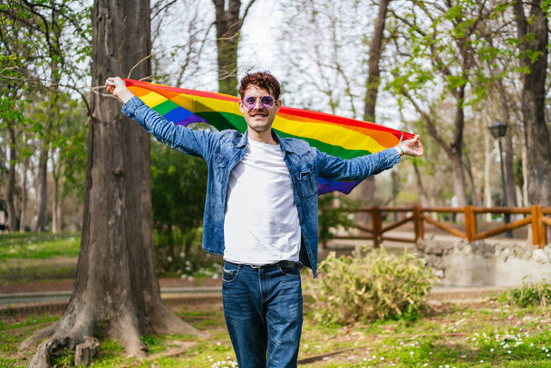 За винятком щастя, гей-чоловік розтягує руки, тримаючи прапор ЛГБТ у яскравому парку. - Фото, зображення