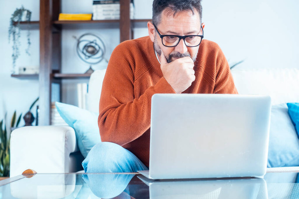 Worried man working from home looking at laptop on sofa. Security problems online lost password concept. Modern mature male people work on computer sitting on couch. Remote worker. Surfing net social - Photo, Image