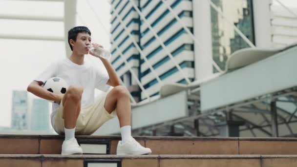 アジアの男性アスリートは,彼の手のボールと外の長いスポーツ練習の後に飲む. 若いフットボール選手が喉が渇いていると感じる水を飲む. サッカーと人々のコンセプト - 映像、動画