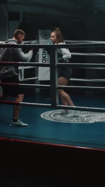 Sportos nő boksz kesztyű készül harci verseny és gyakorlatok férfi edző sötét tornaterem. A női bokszoló bokszkesztyűt üt bokszringen. Függőleges lövés - Felvétel, videó