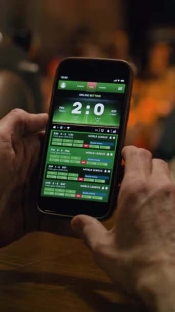 Vertical shot of mature sports fan checking game score and bookmaker ratings in app using mobile phone. Caucasian man bets on football match online sitting at bar counter in pub at night. Gambling - Footage, Video