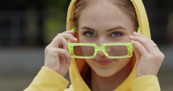 Menina adolescente em capuz amarelo coloca em óculos de sol closeup. A menina ri-se de estar de bom humor durante as férias ao ar livre. Jovem modelo sessão de fotos - Filmagem, Vídeo