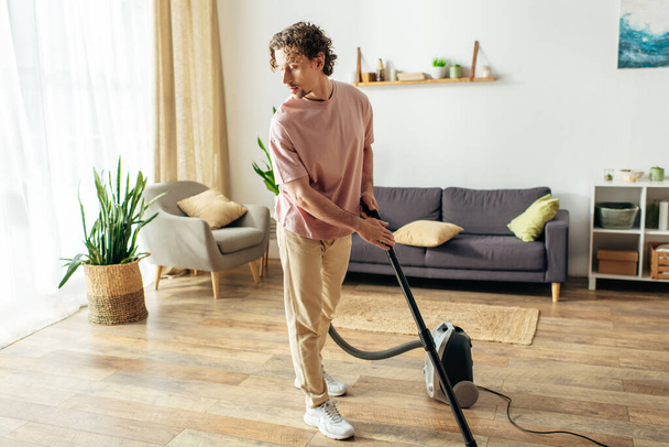 居心地の良いホームウェアのハンサムな男は彼のリビングルームをきれいにするために掃除機を使用します. - 写真・画像