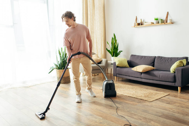 Um homem de roupa de casa aconchegante limpa a sala de estar com um vácuo. - Foto, Imagem
