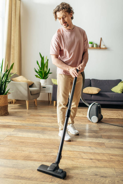 Hombre guapo en ropa de casa acogedora limpia hábilmente el suelo utilizando un vacío. - Foto, Imagen