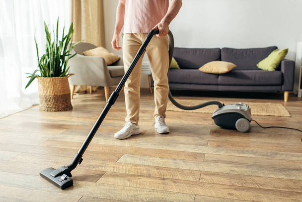 Un hombre guapo en ropa interior acogedora utiliza un vacío para limpiar un piso de madera. - Foto, imagen