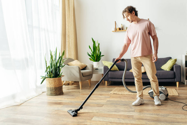 Een knappe man in knusse huiskleding gebruikt een stofzuiger om een woonkamer schoon te maken. - Foto, afbeelding