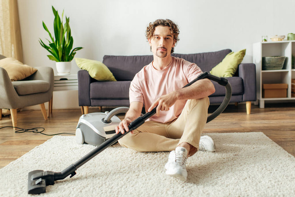 A handsome man in cozy homewear sits on the floor with a vacuum. - Photo, Image