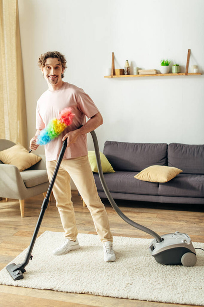 Homem bonito em homewear acolhedor limpa completamente a sala de estar com um aspirador de pó. - Foto, Imagem
