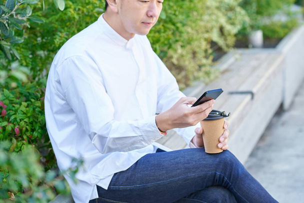 Ένας άνθρωπος που χειρίζεται ένα smartphone πίνοντας καφέ έξω - Φωτογραφία, εικόνα