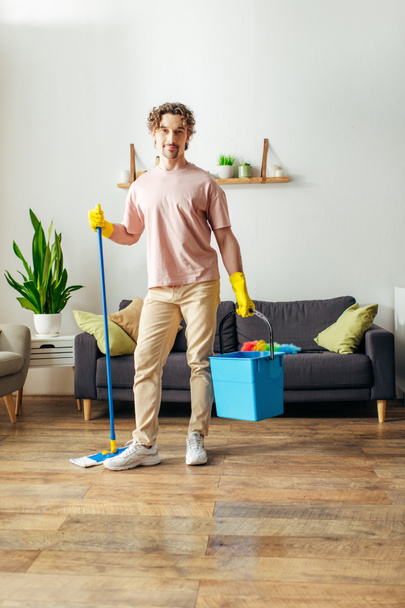 A handsome man in cozy homewear mops the living room floor. - Photo, Image